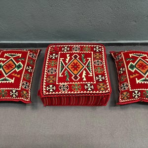 May include: Three red, square cushions with intricate geometric patterns in shades of red, green, and white. The cushions are made of a soft, plush fabric and have a traditional, ethnic design.