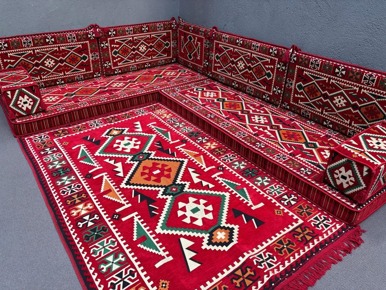 May include: A red and white patterned floor cushion set with a matching rug. The cushions are arranged in a traditional seating configuration, with a large central cushion and smaller cushions on either side. The rug has a geometric design with intricate details.