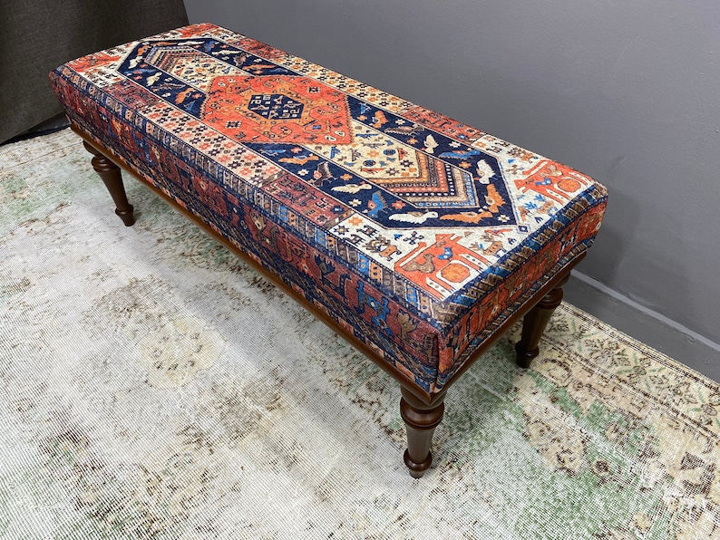 May include: A wooden bench with turned legs and a colourful patterned fabric top. The fabric features a traditional rug design with red, blue, and orange colours.