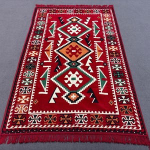 May include: A red rug with a geometric pattern in black, white, green, and orange. The rug has a fringed border.