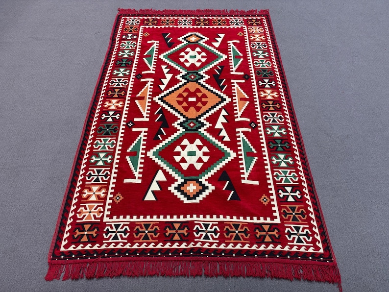 May include: A red rug with a geometric pattern in black, white, green, and orange. The rug has a fringed border.