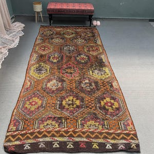 Könnte beinhalten: Ein rechteckiger Teppich mit geometrischem Muster. Der Teppich weist ein sich wiederholendes Muster aus Sechsecken in Orange-, Braun-, Gelb- und Rottönen auf. Der Rand hat eine dunkelbraune Farbe mit einem sich wiederholenden geometrischen Muster. Der Teppich liegt auf einem grauen Boden.