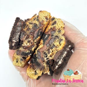 Puede incluir: Una galleta partida por la mitad con una base de masa dorada incrustada con chispas de chocolate oscuro y trozos de galletas negras tipo sándwich con relleno de crema blanca. El interior muestra un centro de chocolate fundido.