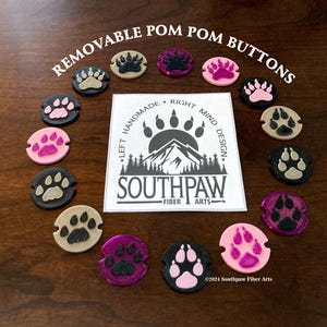 May include: A set of 12 colorful paw print buttons with removable pom poms. The buttons are made of polymer clay and feature a variety of colors, including pink, purple, black, and beige. The buttons are arranged in a circle around a logo that reads "Southpaw Fiber Arts".
