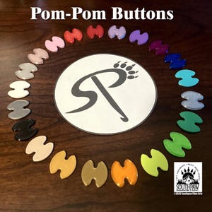 May include: A collection of colorful pom-pom buttons arranged in a circle. The buttons are in various colors, including pink, red, purple, blue, and green. The image also features a circular logo with the text "Pom-Pom Buttons" and the Southpaw Fiber Arts logo.