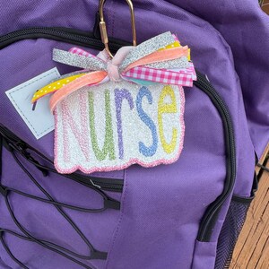 May include: A purple backpack with black zippers and drawstrings. A decorative tag with the word "NURSE" in pastel colors is attached with a gold carabiner clip. The tag has a white background and is adorned with a multi-colored bow.