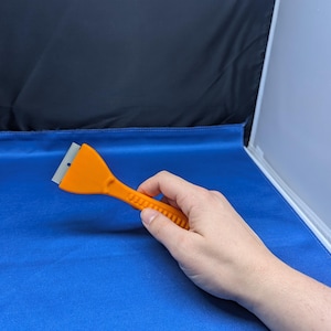 May include: An orange plastic scraper with a metal blade. The scraper is held in a hand.