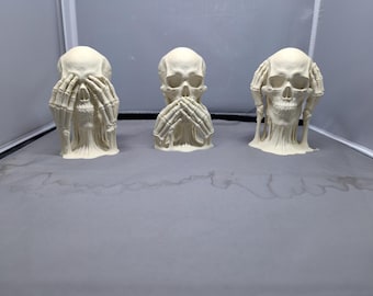 May include: Three off-white skull sculptures, each in a different pose of the "See no evil, Hear no evil, Speak no evil" concept. The skulls have hands covering their eyes, mouth, and ears. The sculptures are on a gray surface.