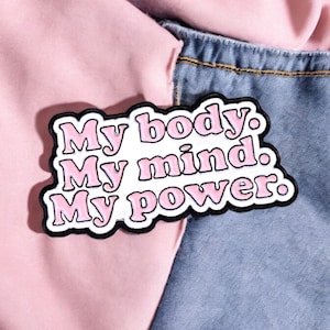 Op de afbeelding: Een roze en witte emaille pin met de woorden "My body. My mind. My power." in een bubble letter lettertype. De pin heeft een zwarte omtrek en is geplaatst tegen een achtergrond van roze stof en denim.