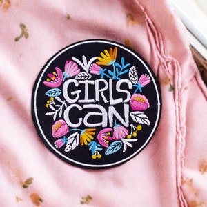 Könnte beinhalten: Ein gestickter Patch in Schwarzweiß mit einem Kreis aus bunten Blumen um den Text "GIRLS CAN".