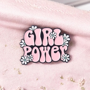 Op de afbeelding: Een roze emaille pin met de woorden "GIRL POWER" in een retro lettertype, omlijnd in zwart. De pin is versierd met kleine witte en lichtblauwe bloemaccenten. De pin is geplaatst tegen een roze stoffen achtergrond.