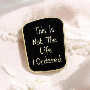 Può includere: Spilla in smalto nero con bordo dorato con il testo "This is not the life I ordered".