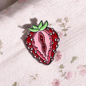 Peut inclure: Une épingle en émail rouge et rose en forme de fraise tranchée. La broche présente un design détaillé avec une tige verte et des graines blanches. L'intérieur est de couleur rose clair. L'épingle est posée sur un fond à motifs floraux.