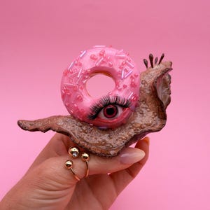 Peut inclure: Une sculpture fantaisiste d'un escargot avec une coquille de donut rose glacé, ornée de vermicelles. Le corps de l'escargot est brun texturé, et son œil est un œil réaliste et ouvert avec de longs cils. Une bague dorée à trois sphères est visible.