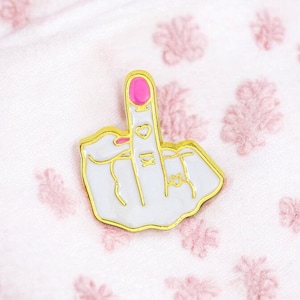 May include: Gold-tone enamel pin featuring a white middle finger with pink nail polish and a heart and a line tattoo.