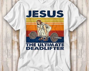May include: White t-shirt with a vintage-style graphic of Jesus lifting weights. The graphic features a retro color palette with orange, yellow, and red stripes. The text reads "JESUS THE ULTIMATE DEADLIFTER".