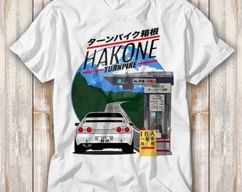 May include: White t-shirt with a graphic of a white car driving on a winding road with mountains in the background. The text "Hakone Turnpike" is printed in red and blue on the shirt. The text "ターンパイク箱根" is printed in black on the shirt.