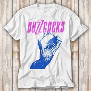 May include: White t-shirt with a blue and pink graphic of a woman with a record player on her head. The text "Buzzcocks" is printed in pink above the graphic.