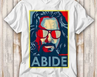May include: A white t-shirt with a red, white, and blue graphic of a man with long hair and sunglasses. The graphic has the word "ABIDE" in yellow letters below it.