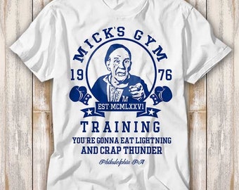 May include: White t-shirt with a blue graphic of a boxing gym called "Mick's Gym" established in 1976. The graphic features a man with a mustache and a boxing glove, and the text "Training You're gonna eat lightning and crap thunder Philadelphia PA".