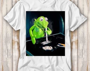 May include: A white t-shirt with a graphic print of a green frog puppet holding a white straw and a pile of white powder on a black surface.