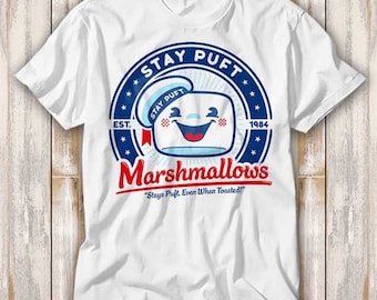 May include: White t-shirt with a blue and red circular graphic featuring a cartoon marshmallow with a sailor hat and the text "Stay Puft Marshmallows" and "Stays Puft, Even When Toasted!"