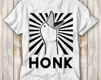 May include: White t-shirt with a black and white illustration of a goose with its mouth open, a black and white striped background, and the word "HONK" in black letters.