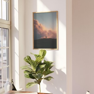 a serene indoor setting with a large window, a framed picture of a sunset, a potted plant, and a wooden table with a book on it.