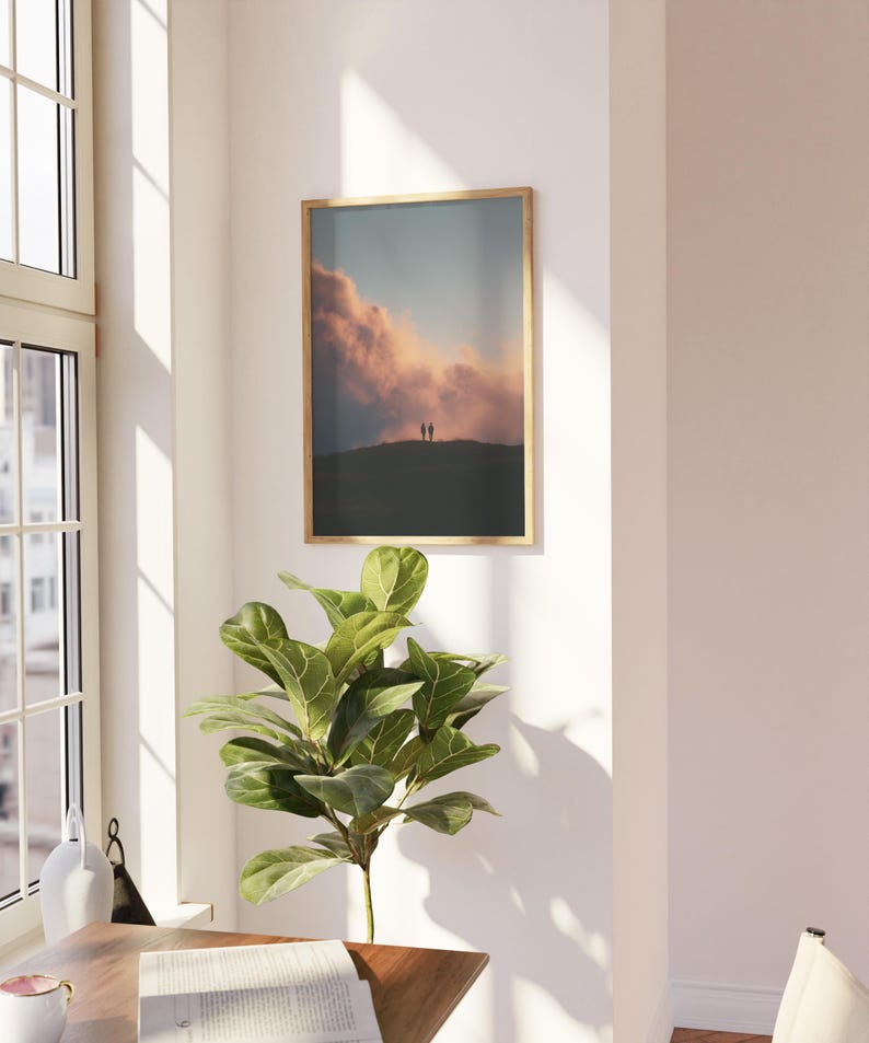 a serene indoor setting with a large window, a framed picture of a sunset, a potted plant, and a wooden table with a book on it.