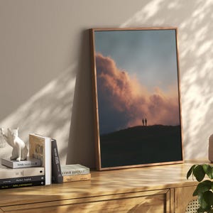 a wooden bookshelf with a framed picture of a sunset over a hill, a stack of books, and a small white figurine of a cat.