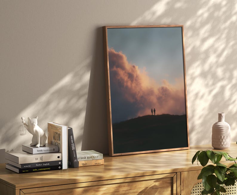 a wooden bookshelf with a framed picture of a sunset over a hill, a stack of books, and a small white figurine of a cat.