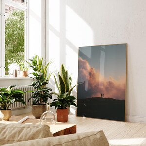 a living room with a large framed picture of a sunset on the wall, a window with plants, and a wooden coffee table.