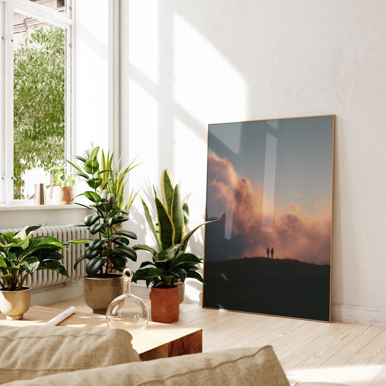 a living room with a large framed picture of a sunset on the wall, a window with plants, and a wooden coffee table.