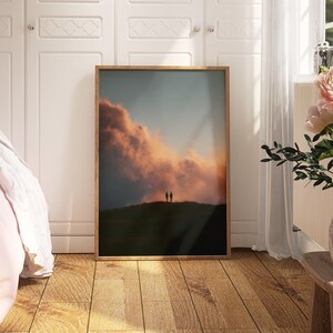 a bedroom with a large framed picture of a sunset on the wall, a vase of pink flowers on a wooden table, and a bed with a white comforter.