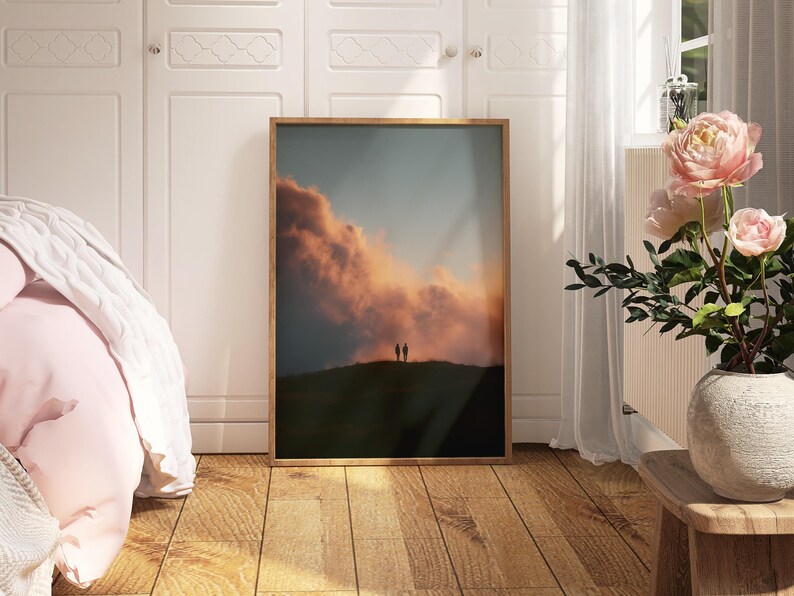 a bedroom with a large framed picture of a sunset on the wall, a vase of pink flowers on a wooden table, and a bed with a white comforter.