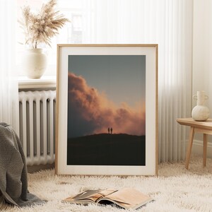 a large framed picture of a sunset over a hill, with two silhouetted figures standing on the hill against the backdrop of the sky. The picture is placed on a white shag rug in a cozy living room setting, with a window, a radiator, and a vase nearby.