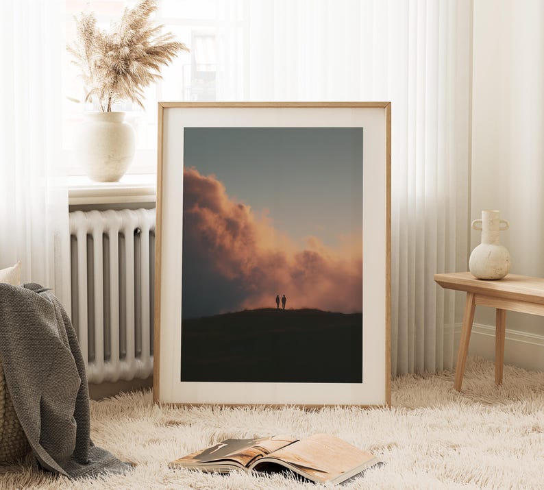 a large framed picture of a sunset over a hill, with two silhouetted figures standing on the hill against the backdrop of the sky. The picture is placed on a white shag rug in a cozy living room setting, with a window, a radiator, and a vase nearby.