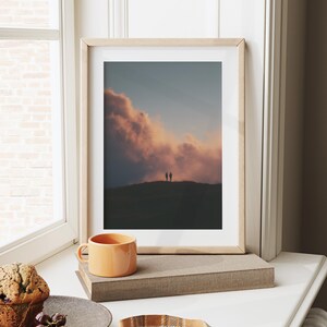 a framed photograph of a couple standing on a hill, with a dramatic sky in the background. The photograph is displayed on a white wall, and there is a cup of coffee and a plate of food nearby.