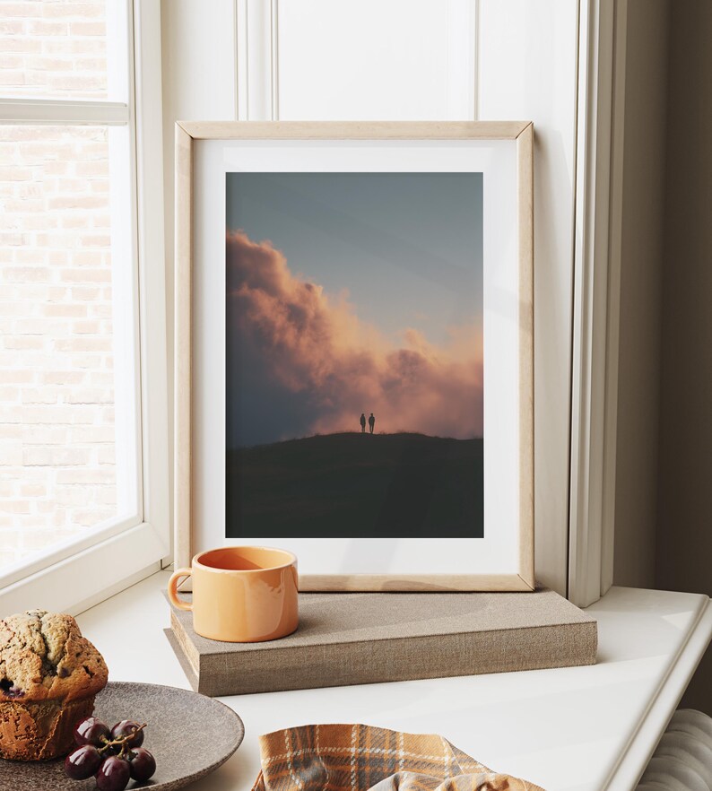 a framed photograph of a couple standing on a hill, with a dramatic sky in the background. The photograph is displayed on a white wall, and there is a cup of coffee and a plate of food nearby.