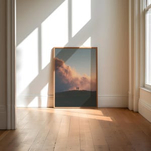 a large framed picture of a sunset over a hill, with two people standing on the hill in the background. The picture is displayed on a wall in a room with a wooden floor.