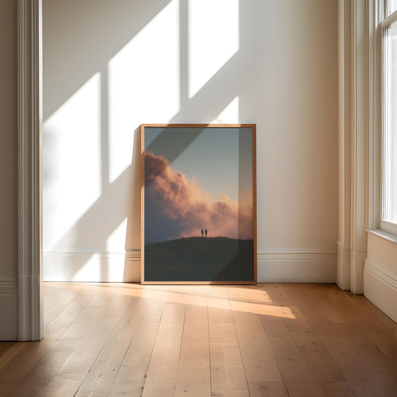 a large framed picture of a sunset over a hill, with two people standing on the hill in the background. The picture is displayed on a wall in a room with a wooden floor.