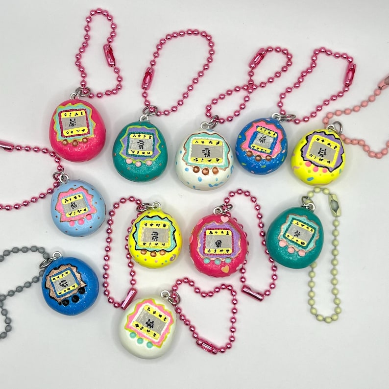 Puede incluir: Una colecci&oacute;n de llaveros Tamagotchi coloridos pintados a mano. Cada llavero presenta un color diferente, incluyendo rosa, azul, verde y amarillo, con una peque&ntilde;a pantalla que muestra personajes pixelados. Los llaveros est&aacute;n sujetos a cadenas de bolas rosas o plateadas.