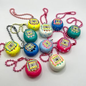 Puede incluir: Una colecci&oacute;n de llaveros coloridos, pintados a mano, con forma de huevo. Cada llavero presenta un peque&ntilde;o dise&ntilde;o de pantalla con texto japon&eacute;s y est&aacute; unido a una cadena de cuentas. Los llaveros son de varios colores, incluyendo amarillo, azul, rosa y verde.