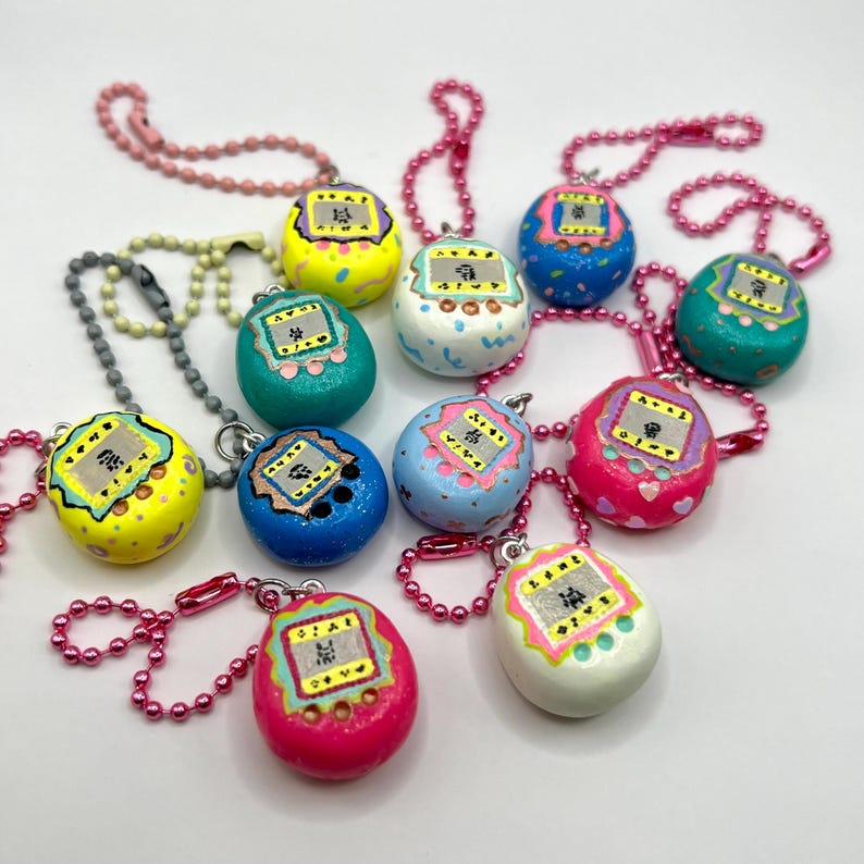 Puede incluir: Una colecci&oacute;n de llaveros coloridos, pintados a mano, con forma de huevo. Cada llavero presenta un peque&ntilde;o dise&ntilde;o de pantalla con texto japon&eacute;s y est&aacute; unido a una cadena de cuentas. Los llaveros son de varios colores, incluyendo amarillo, azul, rosa y verde.