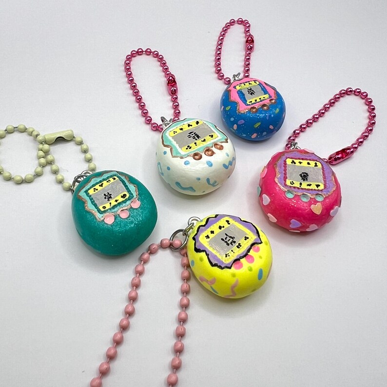 Puede incluir: Una colecci&oacute;n de colgantes coloridos, pintados a mano, con forma de huevo, que recuerdan a los juguetes digitales para mascotas vintage. Cada colgante presenta un peque&ntilde;o dise&ntilde;o de pantalla con caracteres japoneses y est&aacute; unido a una cadena de cuentas rosa o plateada.