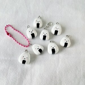 May include: A set of nine white onigiri charms with black seaweed and pink cheeks. The charms are attached to silver chains and some have pink beads.