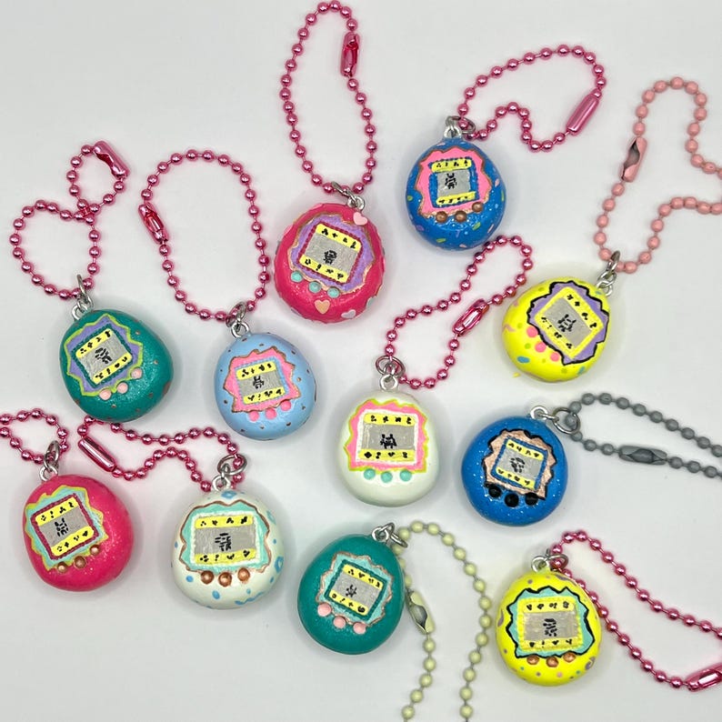 Puede incluir: Una colecci&oacute;n de llaveros coloridos pintados a mano al estilo Tamagotchi. Cada llavero presenta una combinaci&oacute;n de colores &uacute;nica, incluyendo rosa, azul, verde y amarillo, con un peque&ntilde;o dise&ntilde;o de pantalla y una cadena de bolas.