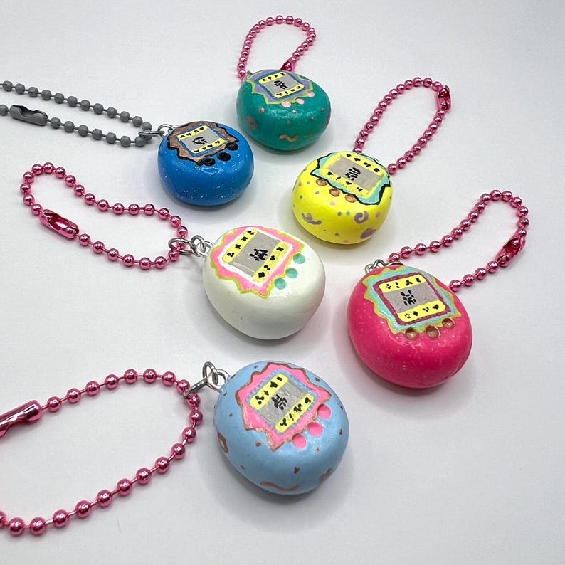 Puede incluir: Una colecci&oacute;n de llaveros coloridos en forma de huevo en azul, verde, amarillo, rosa, blanco y azul claro. Cada llavero presenta un peque&ntilde;o dise&ntilde;o de pantalla con caracteres japoneses y est&aacute; unido a una cadena de cuentas rosa o gris.