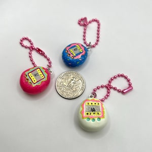 Puede incluir: Tres llaveros de juegos electr&oacute;nicos port&aacute;tiles en miniatura y coloridos. Uno es rosa, otro azul y otro blanco. Cada uno tiene una cadena rosa y una moneda de veinticinco centavos est&aacute; al lado.