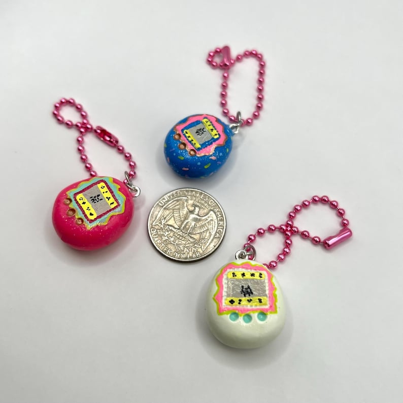 Puede incluir: Tres llaveros de juegos electr&oacute;nicos port&aacute;tiles en miniatura y coloridos. Uno es rosa, otro azul y otro blanco. Cada uno tiene una cadena rosa y una moneda de veinticinco centavos est&aacute; al lado.